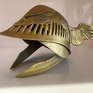 K&ouml;nnte beinhalten: Ein goldener Helm mit gefl&uuml;geltem Design und filigranen Details. Der Helm hat eine spitze Oberseite und einen gebogenen Rand. Die Fl&uuml;gel befinden sich an der Seite des Helms und bestehen aus einem d&uuml;nnen, filigranen Material.