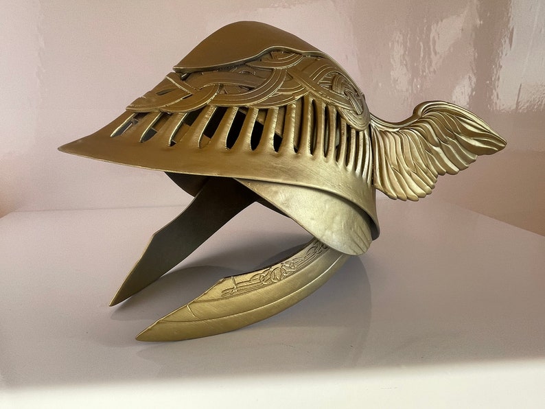 K&ouml;nnte beinhalten: Ein goldener Helm mit gefl&uuml;geltem Design und filigranen Details. Der Helm hat eine spitze Oberseite und einen gebogenen Rand. Die Fl&uuml;gel befinden sich an der Seite des Helms und bestehen aus einem d&uuml;nnen, filigranen Material.