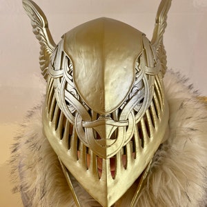 K&ouml;nnte beinhalten: Ein goldfarbener Helm mit aufwendigen Details und gefl&uuml;gelten Akzenten. Der Helm ist aus hartem Material gefertigt und hat eine spitze Oberseite.