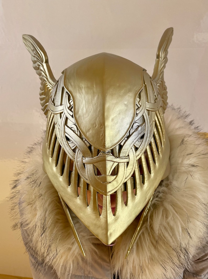 K&ouml;nnte beinhalten: Ein goldfarbener Helm mit aufwendigen Details und gefl&uuml;gelten Akzenten. Der Helm ist aus hartem Material gefertigt und hat eine spitze Oberseite.