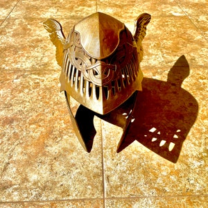 K&ouml;nnte beinhalten: Ein goldfarbener Helm mit filigranen Details und Fl&uuml;geln an den Seiten. Der Helm ist aus Metall und hat eine strukturierte Oberfl&auml;che.
