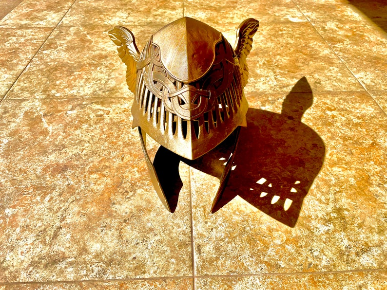 K&ouml;nnte beinhalten: Ein goldfarbener Helm mit filigranen Details und Fl&uuml;geln an den Seiten. Der Helm ist aus Metall und hat eine strukturierte Oberfl&auml;che.