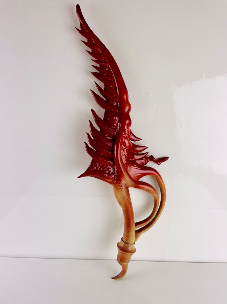 May include: A red and brown decorative sword with a curved blade and a long, thin handle. The blade is shaped like a wing and has a textured surface. The handle is smooth and has a slight curve.