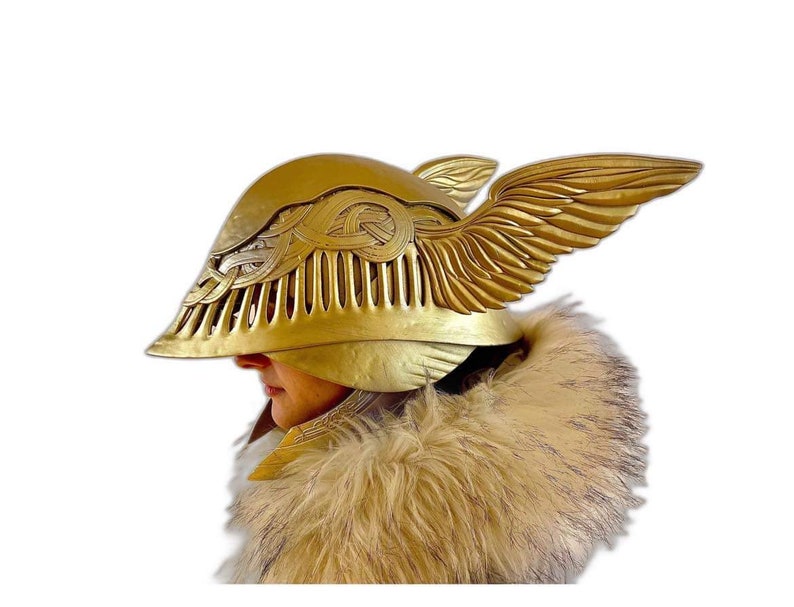 K&ouml;nnte beinhalten: Ein goldener Helm mit Fl&uuml;geldesign. Der Helm hat ein detailliertes, kompliziertes Muster und ein Visier, das das Gesicht bedeckt. Der Helm wird von einer Person mit einem Pelzkragen getragen.