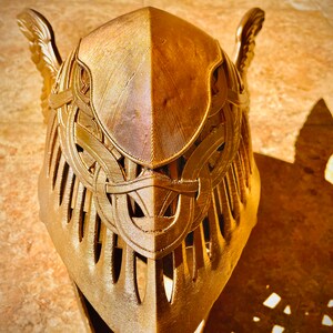 K&ouml;nnte beinhalten: Ein goldfarbener Helm mit komplizierten Details und einem Fl&uuml;geldesign. Der Helm besteht aus einem strukturierten Material und hat eine spitze Oberseite.