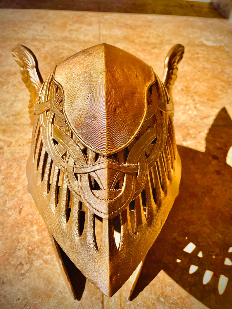K&ouml;nnte beinhalten: Ein goldfarbener Helm mit komplizierten Details und einem Fl&uuml;geldesign. Der Helm besteht aus einem strukturierten Material und hat eine spitze Oberseite.
