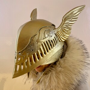 K&ouml;nnte beinhalten: Ein goldener Helm mit Fl&uuml;geln, mit aufwendigen Details und einer strukturierten Oberfl&auml;che. Der Helm ist aus einem harten Material gefertigt und hat eine spitze Form.