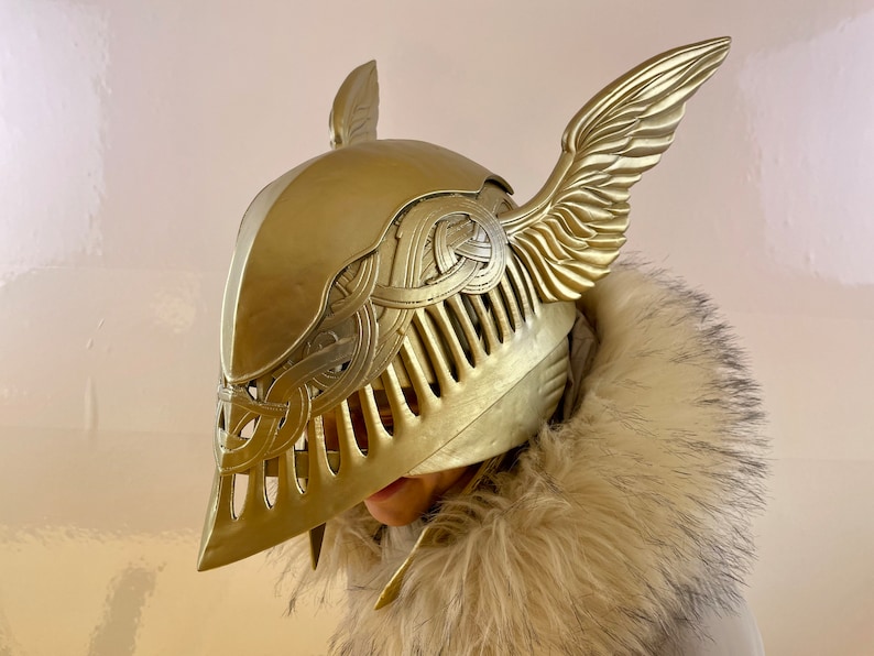 K&ouml;nnte beinhalten: Ein goldener Helm mit Fl&uuml;geln, mit aufwendigen Details und einer strukturierten Oberfl&auml;che. Der Helm ist aus einem harten Material gefertigt und hat eine spitze Form.