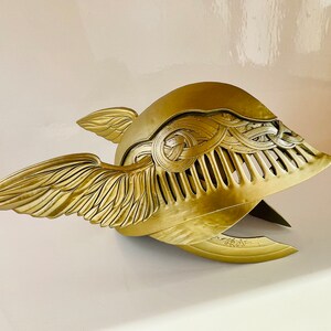 K&ouml;nnte beinhalten: Ein goldener gefl&uuml;gelter Helm mit komplizierten Details und einer strukturierten Oberfl&auml;che. Der Helm hat auf der einen Seite ein Fl&uuml;geldesign und auf der anderen Seite ein dekoratives Muster.