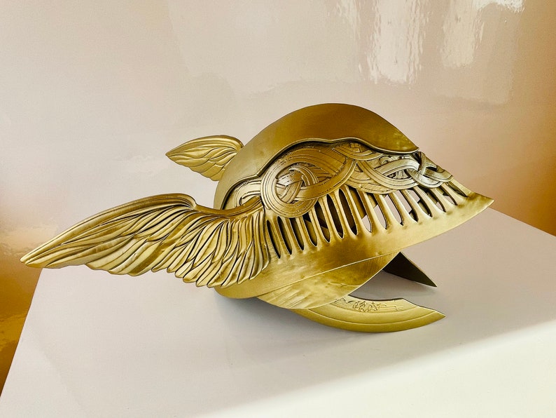 K&ouml;nnte beinhalten: Ein goldener gefl&uuml;gelter Helm mit komplizierten Details und einer strukturierten Oberfl&auml;che. Der Helm hat auf der einen Seite ein Fl&uuml;geldesign und auf der anderen Seite ein dekoratives Muster.