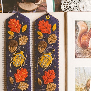 Two felt bookmark featuring the applique of the bug, oak leaves and acorns shown against book spread. The bookmark have knitted acorn charm