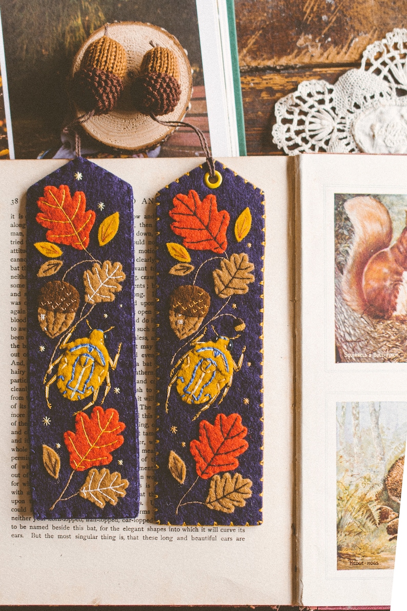 Two felt bookmark featuring the applique of the bug, oak leaves and acorns shown against book spread. The bookmark have knitted acorn charm