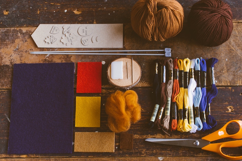 Tools and materials needed to make the bookmark gathered on the dark wood table: felt, needles, scissors, hreads, yarn, pattern and chalk