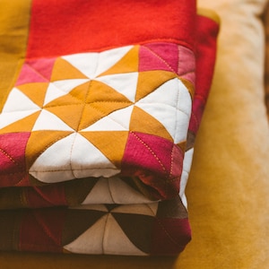 May include: A folded quilt with a patchwork design featuring white, red, and orange triangles. The quilt is folded on top of a yellow surface.
