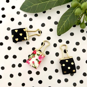 May include: Three binder clips with different patterned fabric covers. The clips are gold and the fabric covers are black with gold hearts, black with gold polka dots, and white with pink flowers.