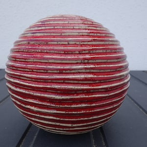 Könnte beinhalten: Ein kugelförmiges Deko-Objekt mit strukturierter Oberfläche. Das Objekt ist überwiegend rot mit weißen horizontalen Streifen. Die runde Form erinnert an einen Globus oder einen großen Ball, geeignet für die Inneneinrichtung.