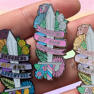 May include: Enamel pins with a sword design, surrounded by flowers and leaves. The pins feature a banner with the text "CHOOSING MY OWN ADVENTURE" in pink and blue. The pins have a silver finish and colorful floral accents.