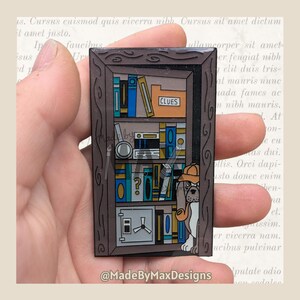 May include: A brown enamel pin shaped like a bookshelf, featuring books, a magnifying glass, and a safe. A dog wearing a detective hat and holding a pipe peeks from behind the shelf. The word "CLUES" is visible on a file folder.