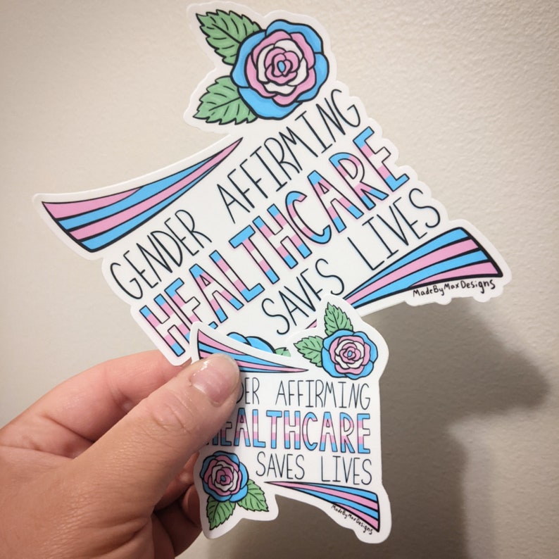 Pu&ograve; includere: Due adesivi con un design a strisce rosa, blu e bianco. Gli adesivi presentano una rosa con foglie verdi e il testo "Gender Affirming Healthcare Saves Lives".