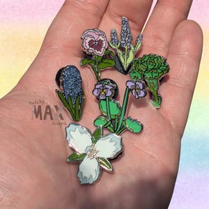 May include: A collection of floral enamel pins in various colors, including purple, blue, green, and white. The pins feature detailed designs of different flowers. The text "Made by MAX designs" is visible.