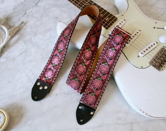 Puede incluir: Una correa de guitarra negra con un patrón floral rosa y verde. La correa tiene extremos de cuero con tachuelas metálicas.