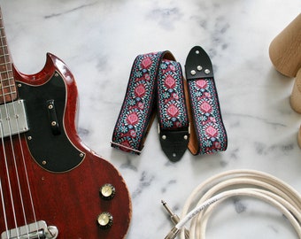 May include: A red electric bass guitar with a black pickguard and a floral patterned guitar strap. The guitar is lying on a white marble surface with a white guitar cable and wooden spools.