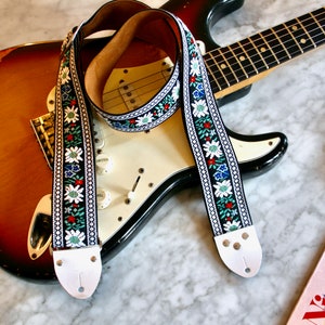 May include: A black guitar strap with white, red, blue, and green floral embroidery. The strap is on a brown and white electric guitar.