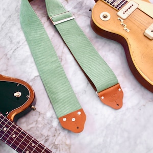 Op de afbeelding: Twee elektrische gitaren met een groene gitaarband. De band heeft een bruin leren uiteinde met de tekst "The Strap Shop" erop.