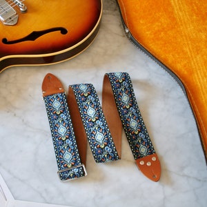 May include: A blue and orange guitar strap with a floral pattern. The strap is made of woven fabric and has a brown leather end.
