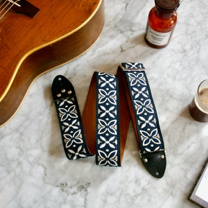 May include: A blue and white patterned guitar strap with leather ends. The strap is lying on a marble surface with a guitar, a bottle, a cup of coffee, and a notebook.