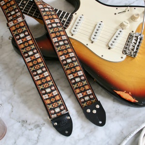 Könnte beinhalten: Ein Gitarrengurt im Vintage-Stil mit schwarzem Hintergrund und einem farbenfrohen geometrischen Muster in Orange, Weiß und Gold. Der Gurt ist an einer Sunburst-E-Gitarre mit weißem Schlagbrett befestigt.