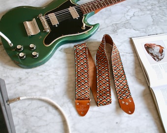 Puede incluir: Una guitarra eléctrica verde con una golpeador negro y una correa de guitarra marrón con un patrón geométrico. La correa tiene un extremo de cuero marrón con el texto "Levy's Leathers" grabado.