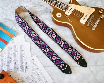 May include: A guitar strap featuring a repeating pattern of purple, pink, and teal geometric shapes on a black background. A gold electric guitar and sheet music are also visible.