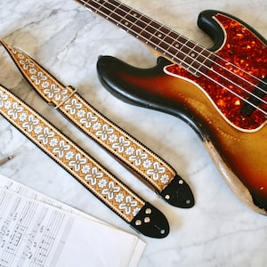 Könnte beinhalten: Ein Gitarrengurt im Vintage-Stil mit einem weißen und goldenen Blumenmuster. Der Gurt ist aus Leder und hat einen Schnallenverschluss. Der Gurt liegt auf einer weißen Marmorfläche neben einer Gitarre und Notenpapier.