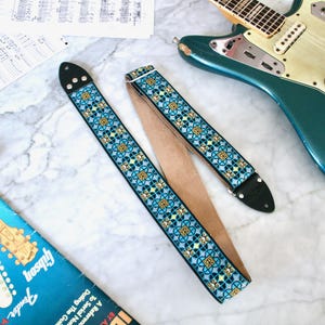 Könnte beinhalten: Ein Gitarrengurt mit einem blauen, gelben und schwarzen geometrischen Muster. Der Gurt besteht aus gewebtem Material mit Lederenden. Eine türkisfarbene E-Gitarre und ein orangefarbenes Effektpedal sind ebenfalls zu sehen.