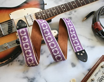 May include: Purple and white guitar strap with a brown leather backing. The strap has a geometric pattern and is shown with a guitar and a guitar amplifier.