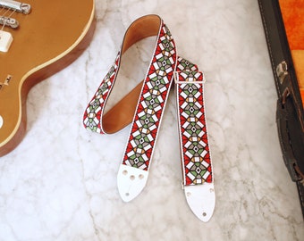 May include: A white guitar strap with a geometric pattern in red, green, and yellow. The strap has white leather ends with metal buckles.