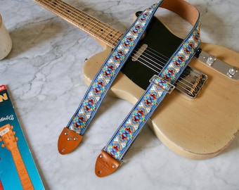 May include: A light blue guitar strap with a colorful geometric pattern. The strap is attached to a light brown electric guitar with a black pickguard.