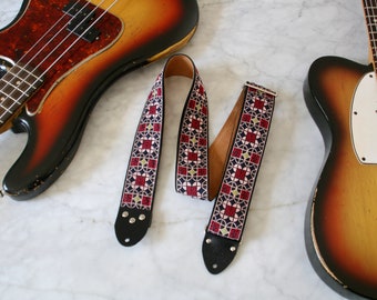 May include: A guitar strap with a geometric pattern in shades of red, pink, blue, and gold. The strap is made of leather and has a black leather end with metal buckles.