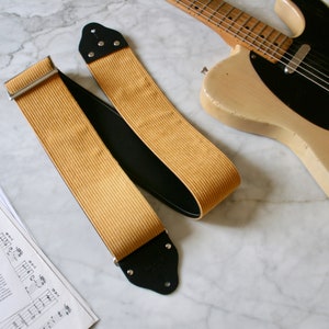 Op de afbeelding: Een gele corduroy gitaarband met zwarte leren uiteinden en zilveren hardware. De band ligt op een wit marmeren oppervlak.