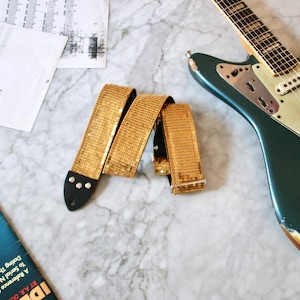 May include: A gold sequined guitar strap with black leather ends. The strap is folded in half and lying on a white marble surface.