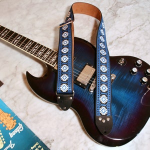 May include: A blue electric guitar with a white and blue patterned guitar strap. The guitar has a black pickguard and a white knob. The guitar is lying on a white marble surface.