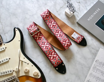 May include: Two red and white patterned guitar straps with brown leather backing. The straps have a black buckle and a black end piece. The straps are lying on a white marble surface next to a white electric guitar.