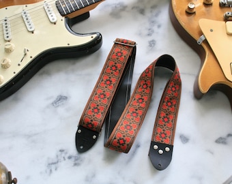 May include: A brown guitar strap with a red and black geometric pattern. The strap has two black leather ends with metal buckles.