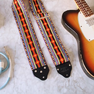 May include: A colorful guitar strap with a geometric pattern in red, orange, yellow, and blue. The strap is paired with a sunburst electric guitar, a light blue cable, and an amplifier.