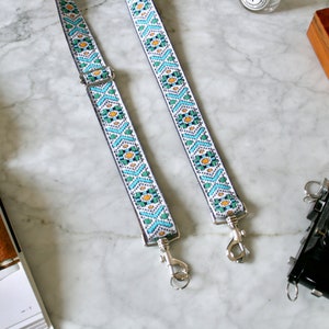 Op de afbeelding: Twee witte camerabandjes met een geometrisch patroon in blauw, groen en geel. De bandjes hebben zilveren metalen gespen.