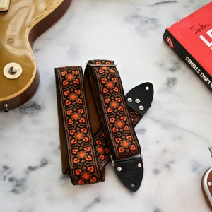 Könnte beinhalten: Ein schwarzer und orangefarbener Gitarrengurt mit geometrischem Muster. Der Gurt hat Lederenden mit Metallnieten.