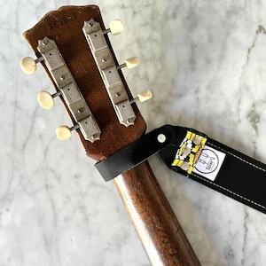 Könnte beinhalten: Nahaufnahme des Kopfes einer braunen Akustikgitarre mit einem schwarzen Ledergitarrengurt. Die Gitarre hat die Seriennummer "283843" und weiße Stimmwirbel. Der Gurt hat ein gelbes und schwarzes Muster mit dem Text "Tom's" darauf.