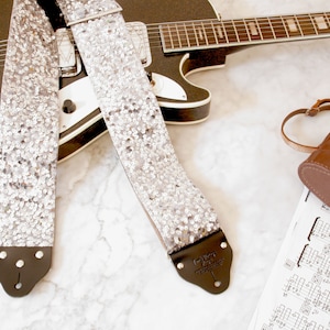 Op de afbeelding: Een zilveren gitaarband met pailletten en zwarte leren uiteinden. De band ligt op een wit marmeren oppervlak met een zwarte elektrische gitaar en een bruine leren cameratas.