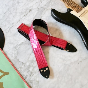 May include: A pink guitar strap with white sequins and a black leather trim. The strap is lying on a white marble surface.